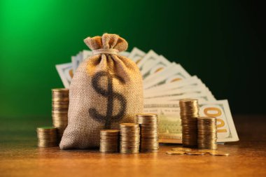Çanta, dolar banknotları ve ahşap masaya yığılmış bozuk paralar yeşil eğimli arka plana karşı