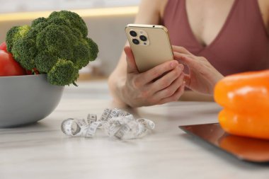Beyaz mermer masada sebzeli akıllı telefon kullanan bir kadın, mutfak terazisi ve kapalı alanda mezura, yakın plan.