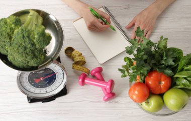 Hafif ahşap masada sağlıklı yiyecekler, mutfak terazisi, mezura ve dambıllarla beslenme planı geliştiren bir kadın.