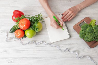 Hafif ahşap masada diyet planı geliştiren kadın mutfak tartısı, mezura ve sağlıklı yiyecekler, üst görünüm.