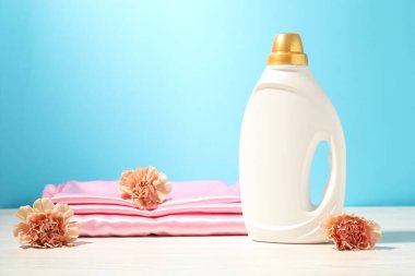 Bir şişe yumuşatıcı, temiz kıyafetler ve karanfil çiçekleri açık mavi arka planda beyaz ahşap masa üzerinde