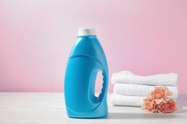 Bir şişe yumuşatıcı, havlular ve karanfil çiçekleri beyaz ahşap masa üzerinde pembe arka planda.