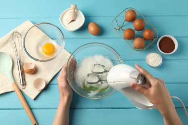 Bir kadın açık mavi ahşap masada el çırpıcı ile yumurta beyazı çırpıyor.