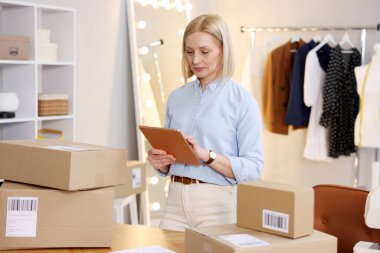 Dükkan sahibi, terzi dükkanında paketlerle birlikte masanın yanında tablet kullanıyor.