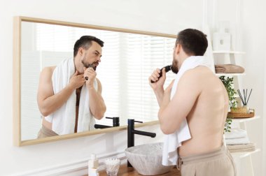 Yakışıklı adam banyoda aynanın yanında tıraş makinesiyle tıraş oluyor.