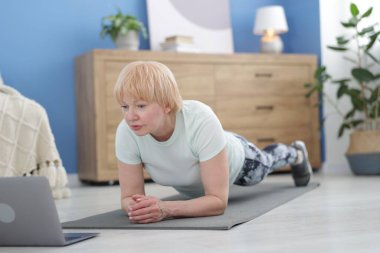 Spor giyim bölümünde son sınıf öğrencisi bir kadın, evde koçla çevrimiçi derste kalas egzersizi yapıyor.