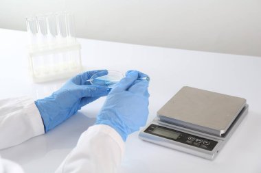 Bilim adamı, beyaz masadaki terazilerin yakınında numuneyle çalışıyor.