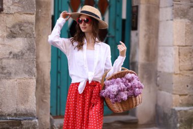 Şapkalı ve güneş gözlüklü güzel bir kadın, bina yakınlarında bir sepet leylak çiçekli.