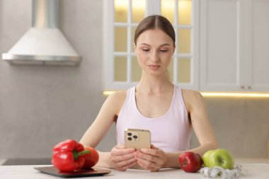 Beyaz masada akıllı telefon kullanan bir kadın. Mutfak terazisi, sağlıklı yiyecekler ve kapalı alanda mezura kullanıyor.