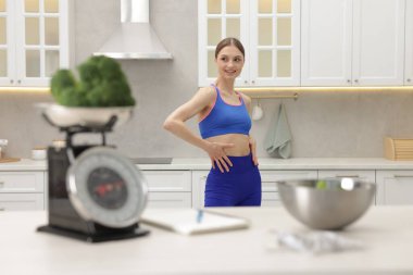 Mutfak terazisi, brokoli, defter, mezura ve salata ile masanın yanında poz veren kadın.