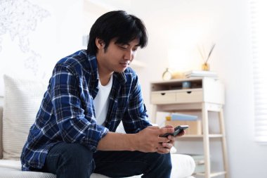 Evdeki kanepede akıllı telefonlu yakışıklı Asyalı adam.