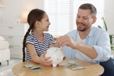 Baba ve kızı kapalı bir masadaki kumbaraya cep harçlığı koyuyor.