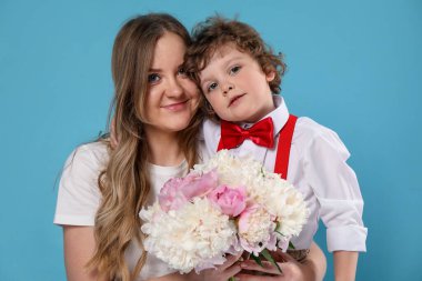 Küçük çocuk annesine açık mavi arka planda şakayık çiçekleri veriyor.