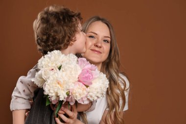 Küçük çocuk annesini öpüyor ve ona kahverengi arka planda şakayık çiçekleri veriyor.