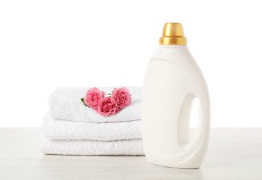 Bir şişe yumuşatıcı, beyaz arka plana karşı ahşap masa üzerine yığılmış havlular ve çiçekler.