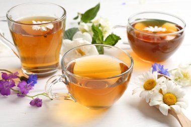 Cam bardaklarda romatik bitki çayı ve beyaz ahşap masada farklı çiçekler, yakın plan.