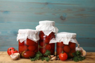 Kavanozlarda domates turşusu taze ürünler ve ahşap masada baharatlar açık mavi arka planda.