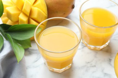 Bardakta lezzetli mango suyu, taze meyve ve yeşil yapraklar beyaz mermer masada, yakın plan.