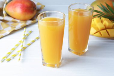 Lezzetli mango suyu bardaklarda, taze meyveler, pipetler ve yeşil yapraklar beyaz ahşap masada, yakın plan