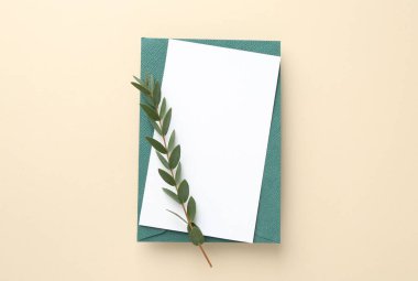 Boş davetiye kartı, zarf ve bej arka planda yeşil dal, üst görünüm