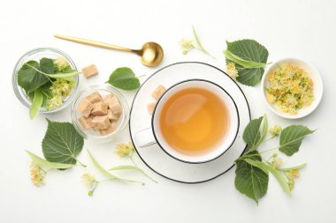 Fincanda tatlı ıhlamur çayı, kaşık, küp şeker, yapraklar ve çiçekler beyaz masada, düz yerde.