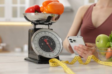 Beyaz mermer masanın yanında akıllı telefon kullanan bir kadın. Mutfak terazisi, sağlıklı yiyecekler ve kapalı alanda mezura kullanıyor.