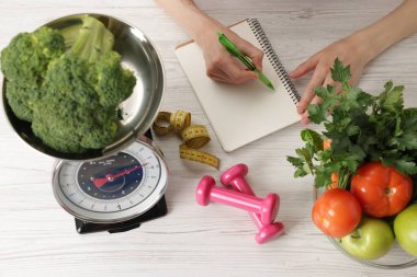 Hafif ahşap masada sağlıklı yiyecekler, mutfak terazisi, mezura ve dambıllarla beslenme planı geliştiren bir kadın.