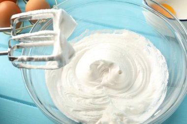 Krem şantili kase, el çırpıcı ve malzemeler açık mavi ahşap masada, yakın plan.