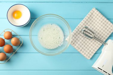 Krem şanti yapıyorum. Kase, el çırpıcı ve malzemeler açık mavi ahşap masada, düz yerde.