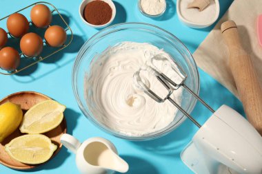 Açık mavi masada çırpılmış krema, yumurta ve diğer ürünlerle el çırpıcı, düz konum