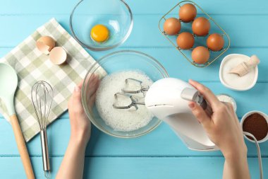 Bir kadın açık mavi ahşap masada el çırpıcı ile yumurta beyazı çırpıyor.