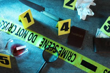 Adli tıp uzmanı. Sarı suç mahalli bandı, işaretler ve kanıtlar gri masada, üst tarafta.