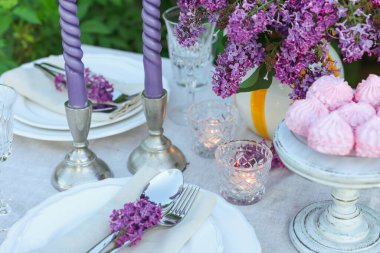 Güzel leylak çiçekleri, zephyrler, tabaklar, çatal bıçak takımı, cam ve mumlar bahçede, yakın plan