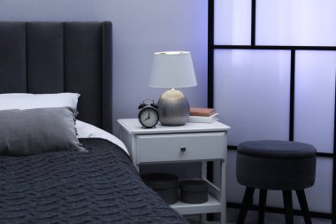 İçinde lamba, çalar saat ve yatağın yanında kitaplar olan komodin.