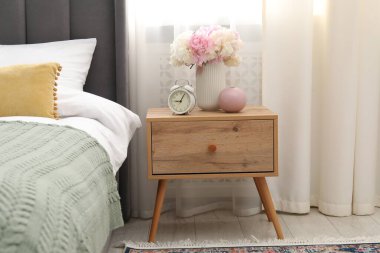 Komodinin yanında çalar saat ve yatağın yanında çiçekler var.