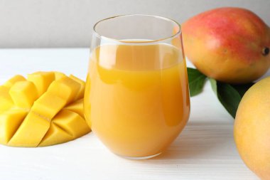 Lezzetli mango suyu bardakta, taze meyve ve yeşil yapraklar beyaz ahşap masada, yakın plan