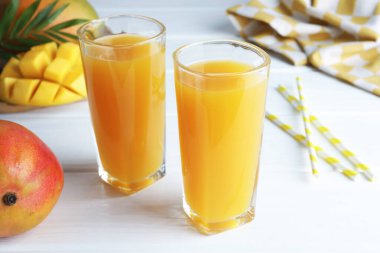 Lezzetli mango suyu bardaklarda, taze meyveler, pipetler ve yeşil yapraklar beyaz ahşap masada, yakın plan