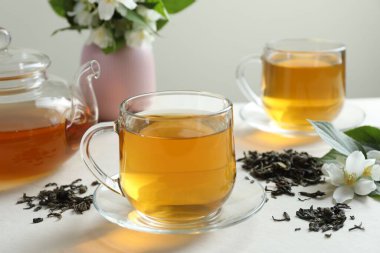 Cam bardaklarda, çaydanlıkta, çiçeklerde ve beyaz masada kurumuş yapraklarda Aromatik yasemin çayı.