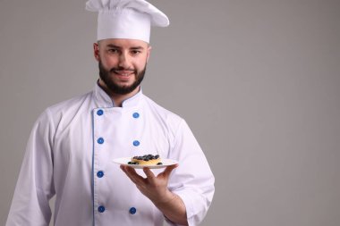 Üniformalı mutlu pastacı açık gri arka planda yaban mersinli lezzetli tart tutuyor.