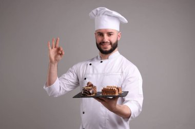 Üniformalı mutlu pastacı lezzetli çikolatalı tatlıları tutuyor ve açık gri arka planda iyi bir jest yapıyor.