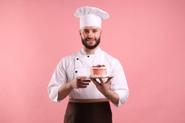 Üniformalı mutlu pastacı pembe arka planda böğürtlenli pasta tutuyor.