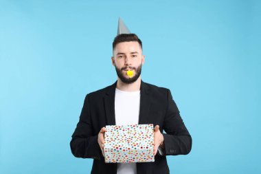 Doğum günün kutlu olsun. Parti şapkası, üflemeli ve açık mavi arka planda hediye kutusu olan yakışıklı bir adam.