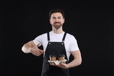 Gülümseyen baristanın portresi siyah arka planda hazır kahve bardaklarına işaret ediyor.