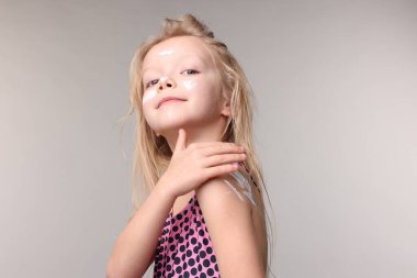 Gri arka planda omzuna güneş kremi süren sevimli küçük bir kız.