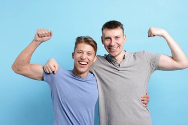 İki arkadaş kucaklaşıp, açık mavi arka planda pazılarını gösteriyorlar.