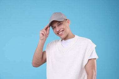 Genç adam açık mavi arka planda şık gri beyzbol şapkası giyiyor.