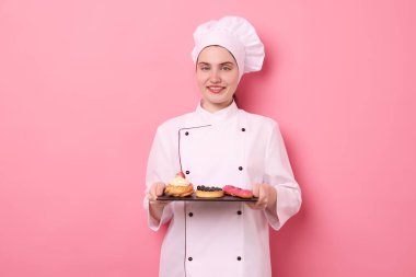 Üniformalı şekerci pembe arka planda farklı lezzetli tatlılar tutuyor.