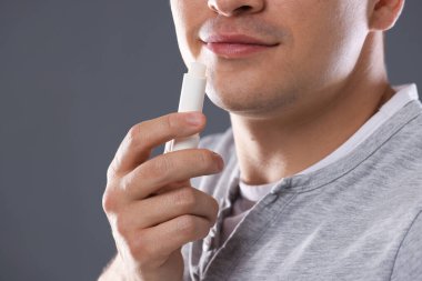 Gri arka planda dudaklarına dudak kremi süren genç adam, yakın plan.