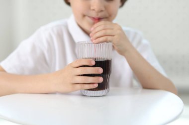 Küçük çocuk, masada ferahlatıcı gazoz içiyor. Kapalı bir yerde.
