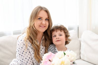 Küçük çocuk evdeki kanepede annesine güzel şakayık çiçekleri veriyor.